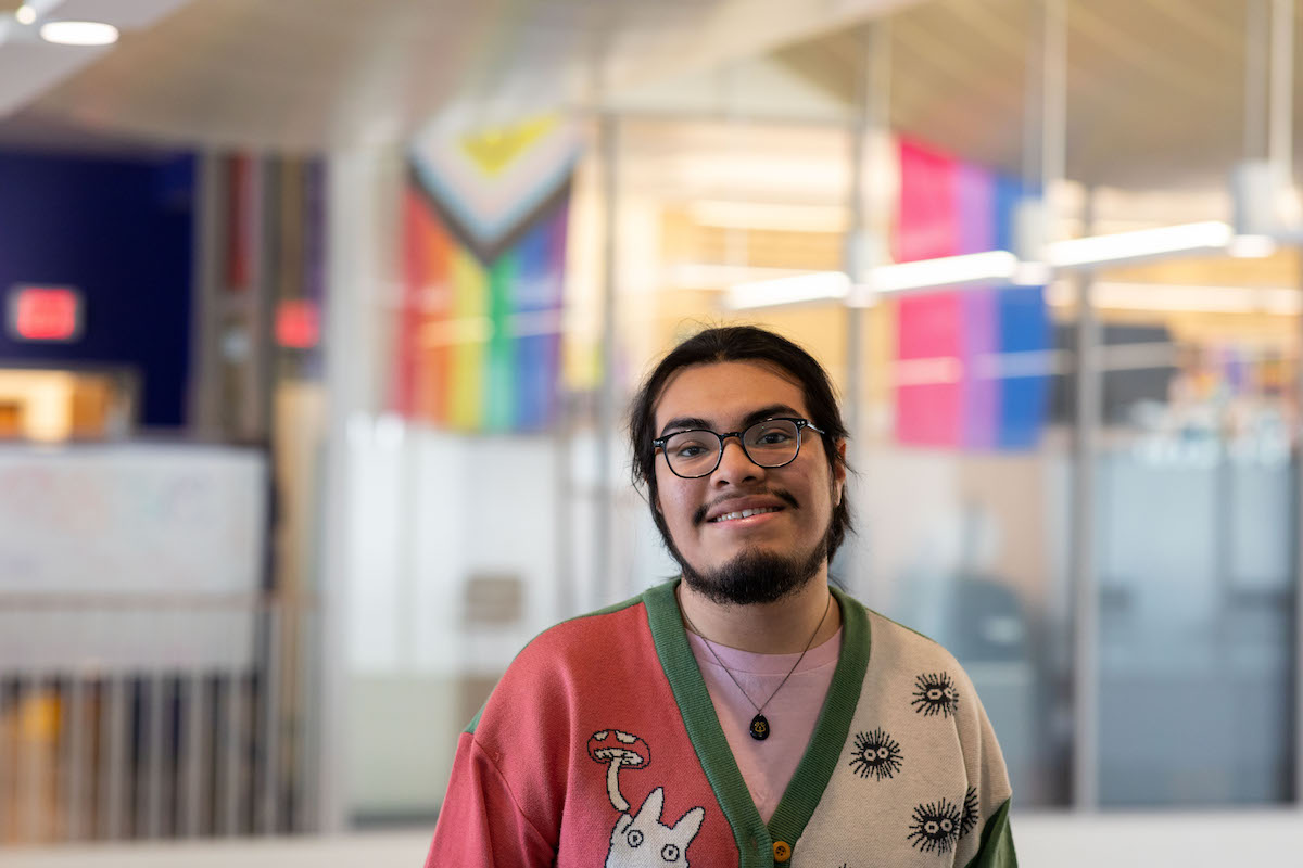 A young person in glasses, a pink shirt and cardigan sweater poses for a portrait in front of a Pride Flag.