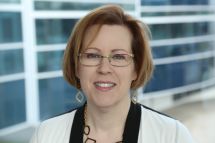 A portrait of a smiling woman with glasses and wearing a white jacket. 