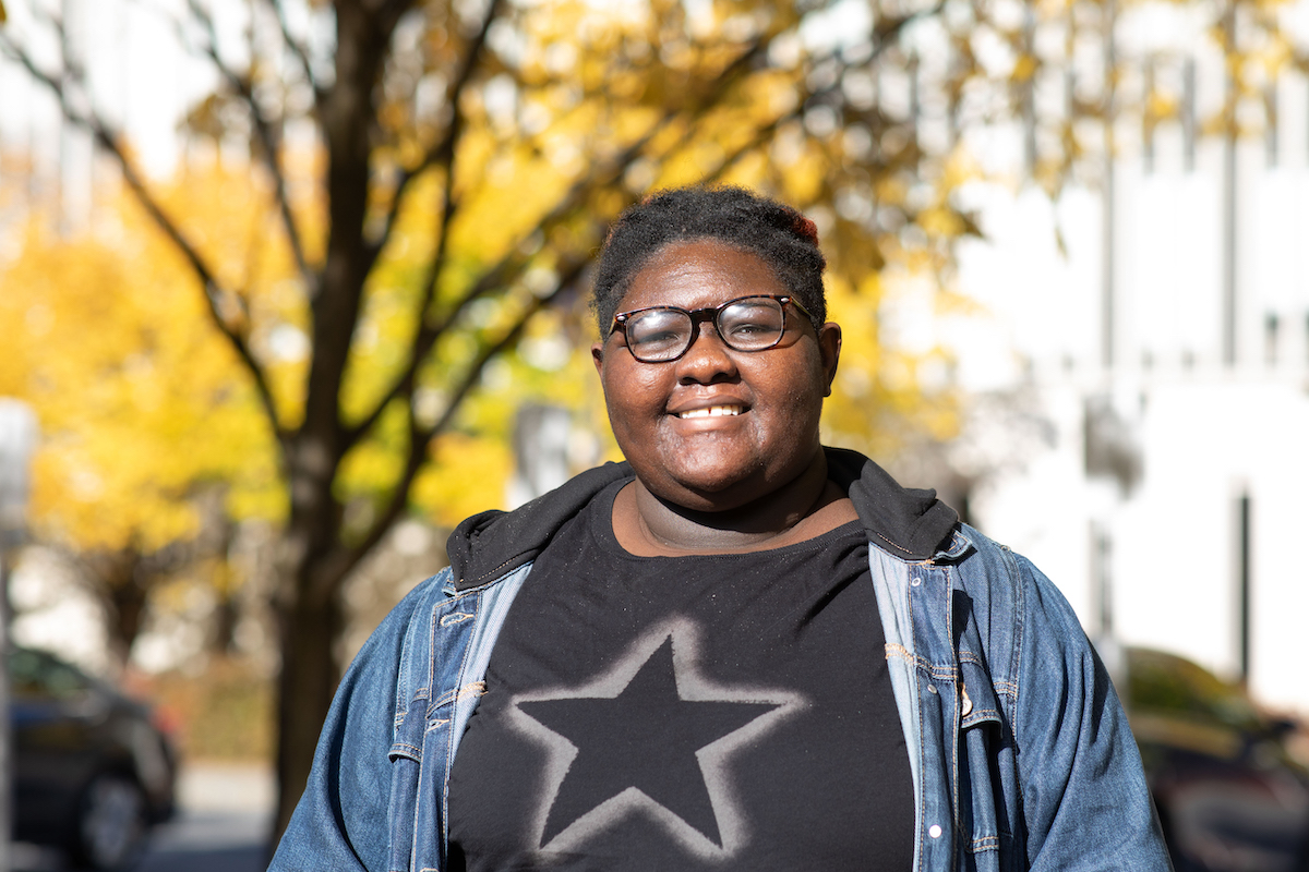 A young person in glasses, a black shirt featuring a star and jean jacket poses outside on a sunny autumn day.