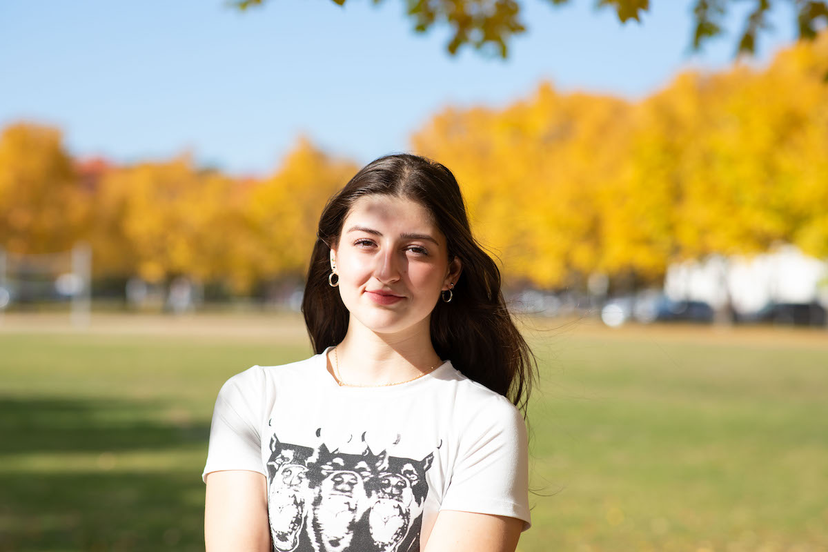 A young person with long brown hair poses for a portrait on campus on a sunny autumn day. A stand of trees with yellow leaves can be seen in the background.