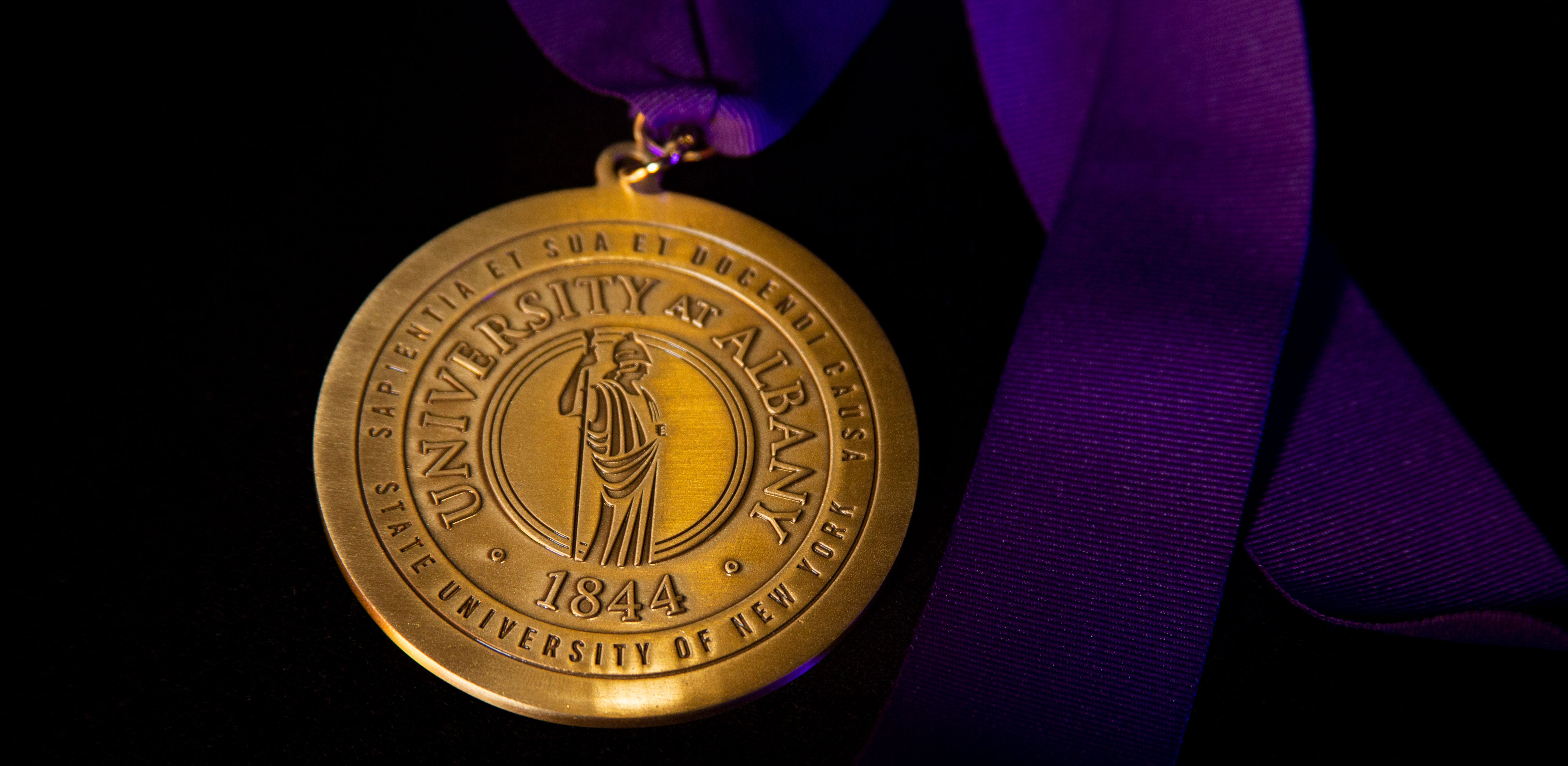 A golden medallion on a purple silk ribbon. The medallion includes the Greek goddess of knowledge, Minerva, and the words, "University at Albany, 1844, Sapientia et sua et docendi causa, State University of New York."