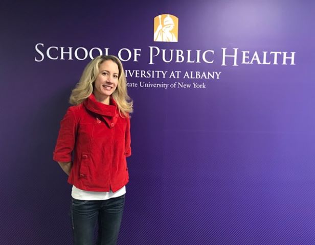 Meg V. stands against a purple wall with the School of Public Health logo on it. She is wearing a red sweater and blue jeans and is smiling at the camera.