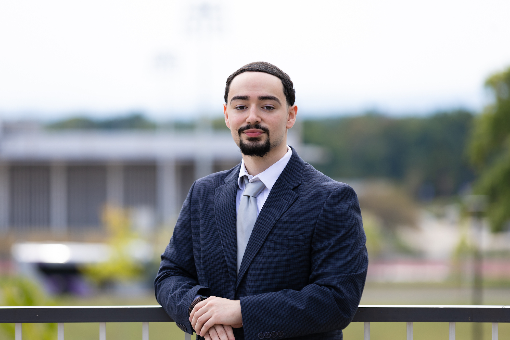 A young man with short black hair and a goatee wearing a blue suit and silver tie leans against a railing outside on the UAlbany campus.