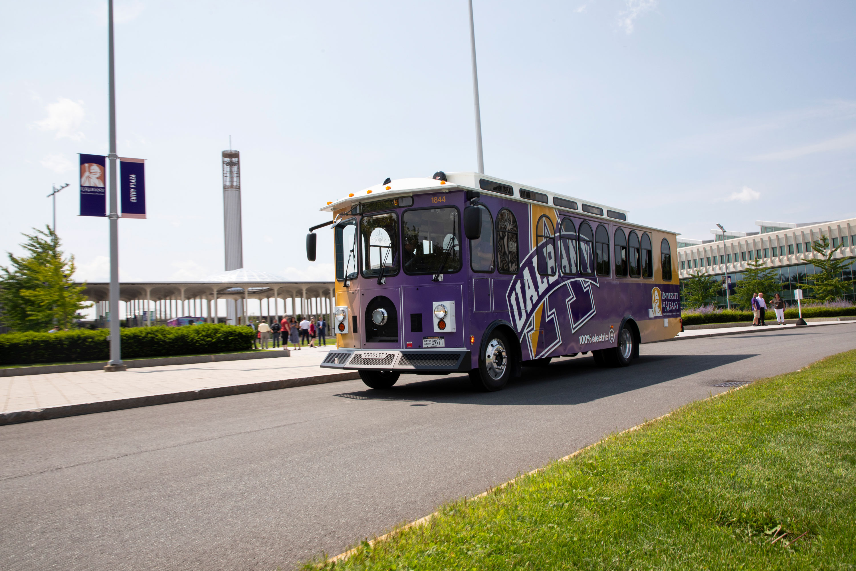 An electric trolley decorated with UAlbany logos and colors parked on Collins Circle on the Uptown Campus, with the carillon in the background.