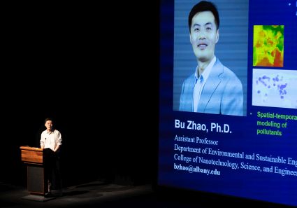 A man stands at a podium on a dark stage next to a projection of his photo and the words Bu Zhao, PhD