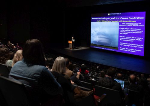 from the back of a theater, the audience can be seen viewing a man at a podium in front of a screen with a picture of a storm and text.