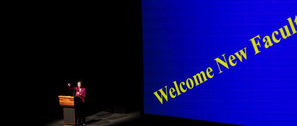 A woman gestures from a podium beside a large blue screen with the words "Welcome New Faculty" in yellow letters.