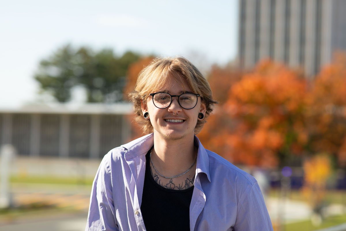 A young person in glasses poses for a portrait on campus on a sunny autumn day.