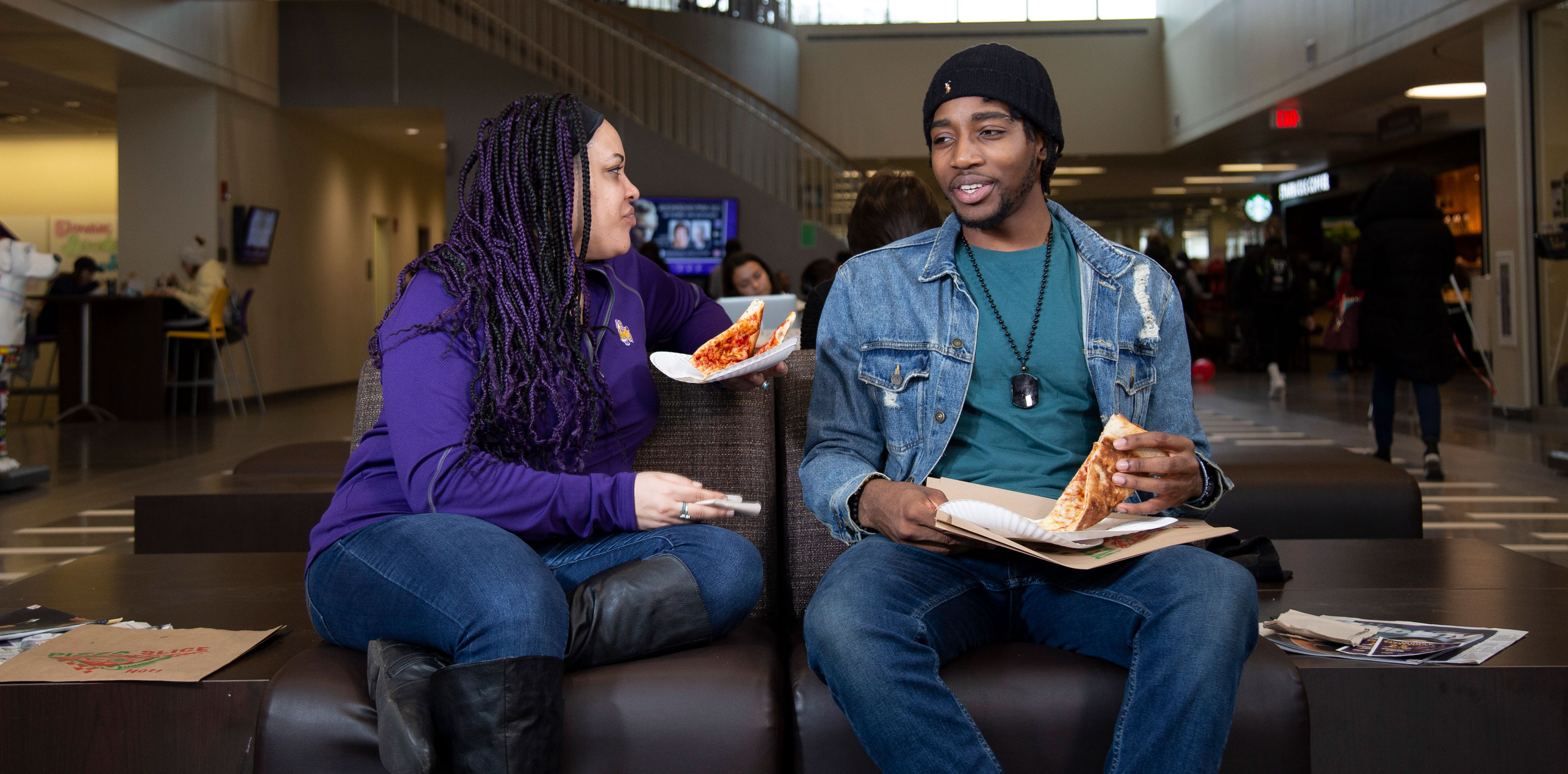 Two students sit on a couch inside Campus Center eating pizza.