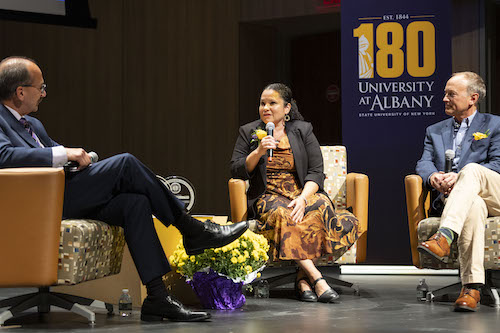 Two men and a woman are seated on stage and have a conversation. A purple banner in the background reads "180 University at Albany"