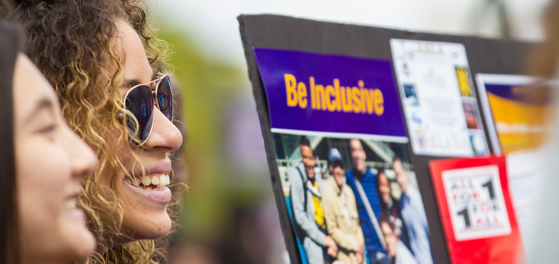 Two women in foreground look in the direction of a sign that reads "Be Inclusive."