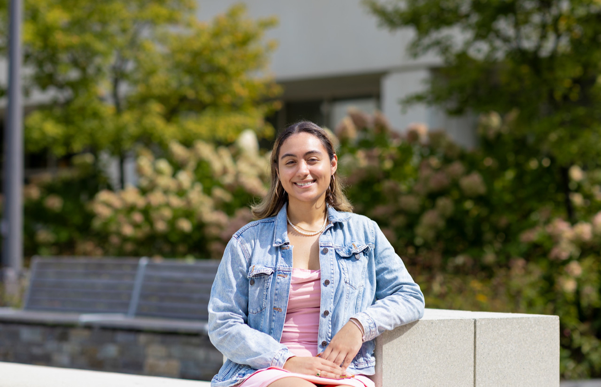 A young woman with shoulder-length brown hair wearing a denim jacket over a pink dress sits on a bench on a sunny day.