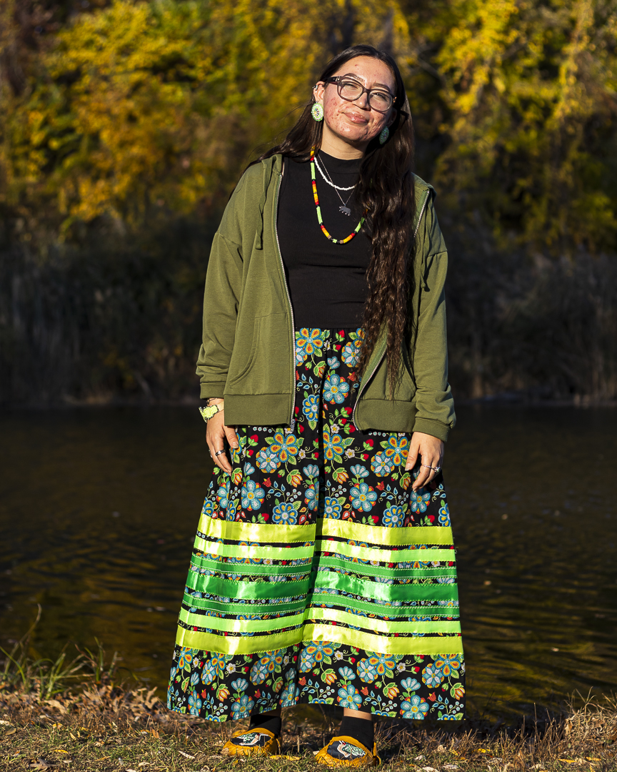 A young woman with long black hair and glasses wears a green sweater over a long colorful skirt and poses for a portrait outdoors on a sunny fall day.