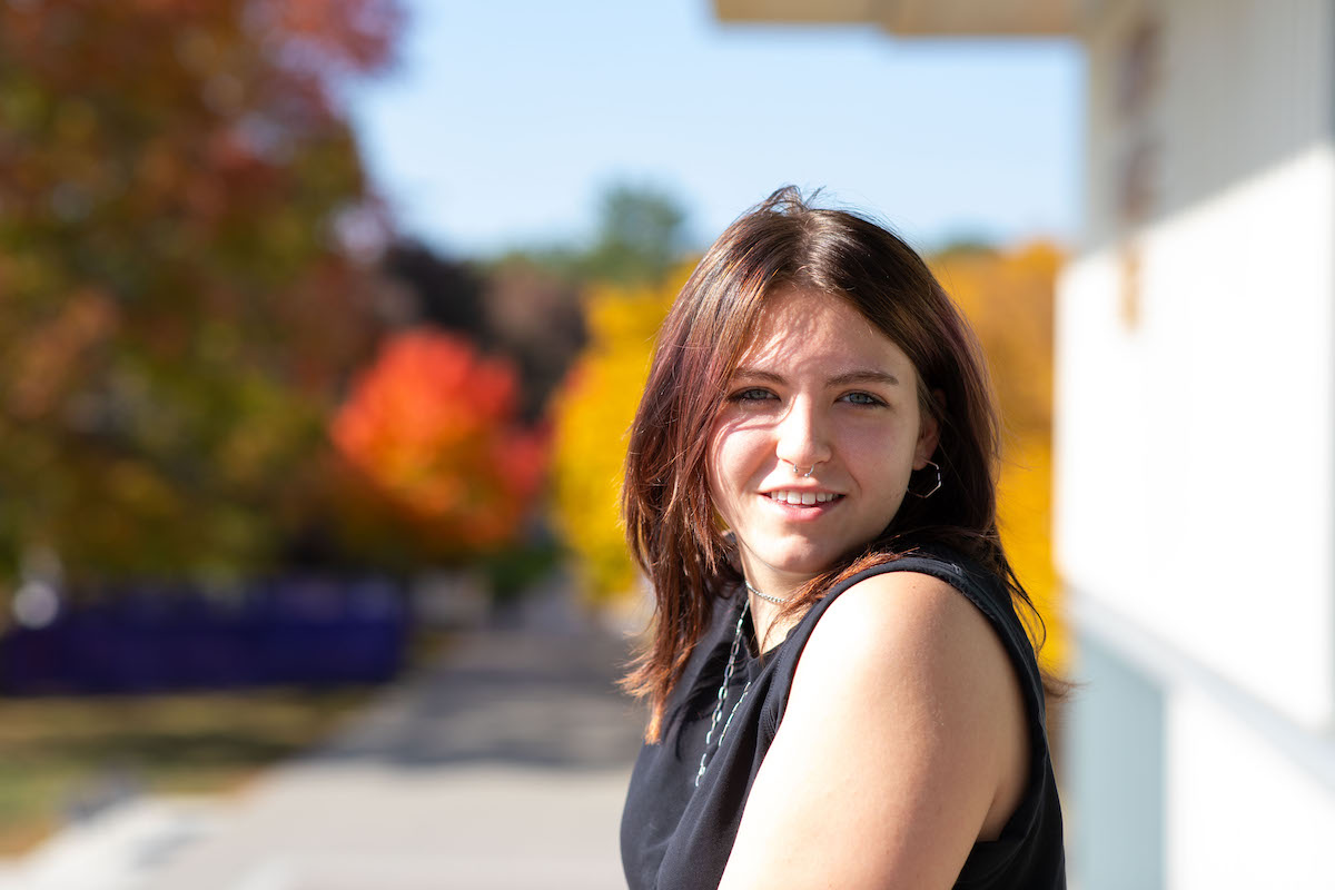 A young person with long brown hair in a black shirt poses for a portrait on a sunny autumn day. Colorful foliage can be seen in the background.