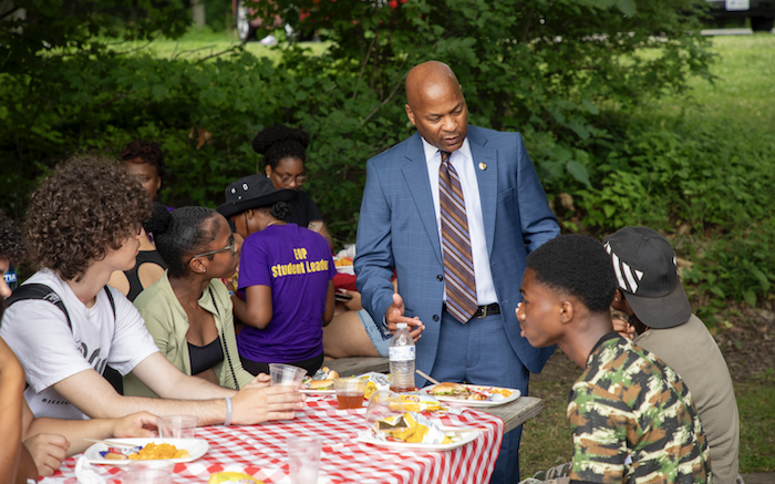 A man in a blue suit speaks to students seated at a picnic table in the woods.
