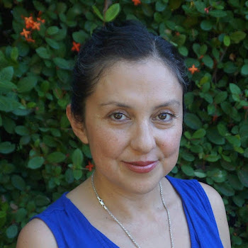 A smiling woman in a blue top poses in front of a green bush.