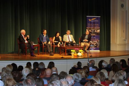 5 people sit on a stage in front of a black curtain; the heads of audience members are seen in front of the stage