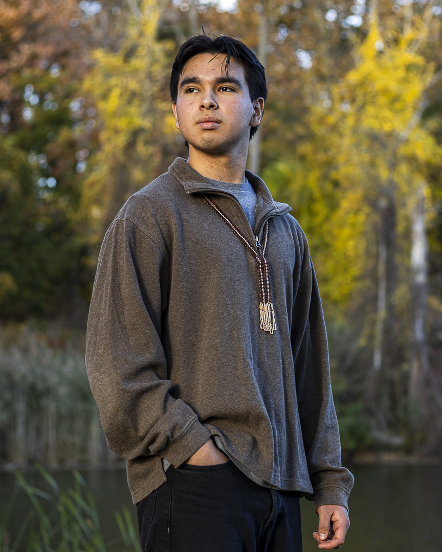 A young man with short black hair wears a gray sweater and poses for a portrat outdoors on a sunny fall day.