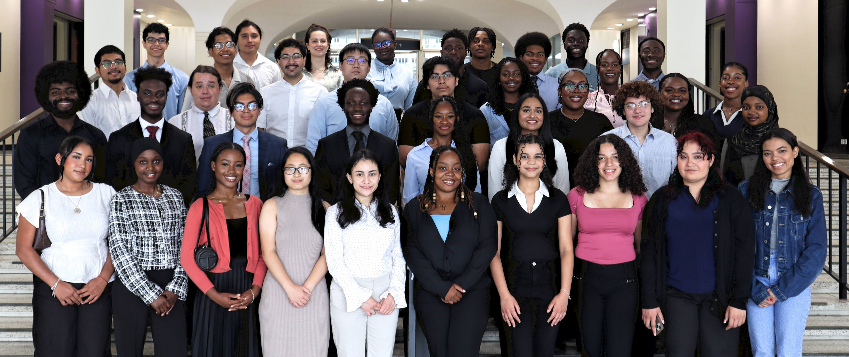 About three dozen of UAlbany's Summer Research Program participants wearing business casual clothing pose for a photo on an indoor staircase on campus.