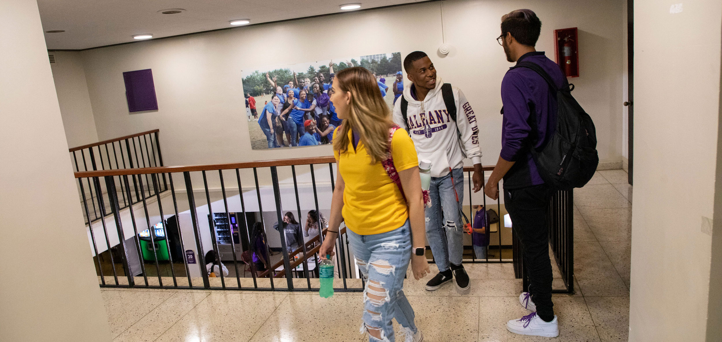 Three students walk by a stairwell inside a residence hall. Vending machines are visible downstairs.