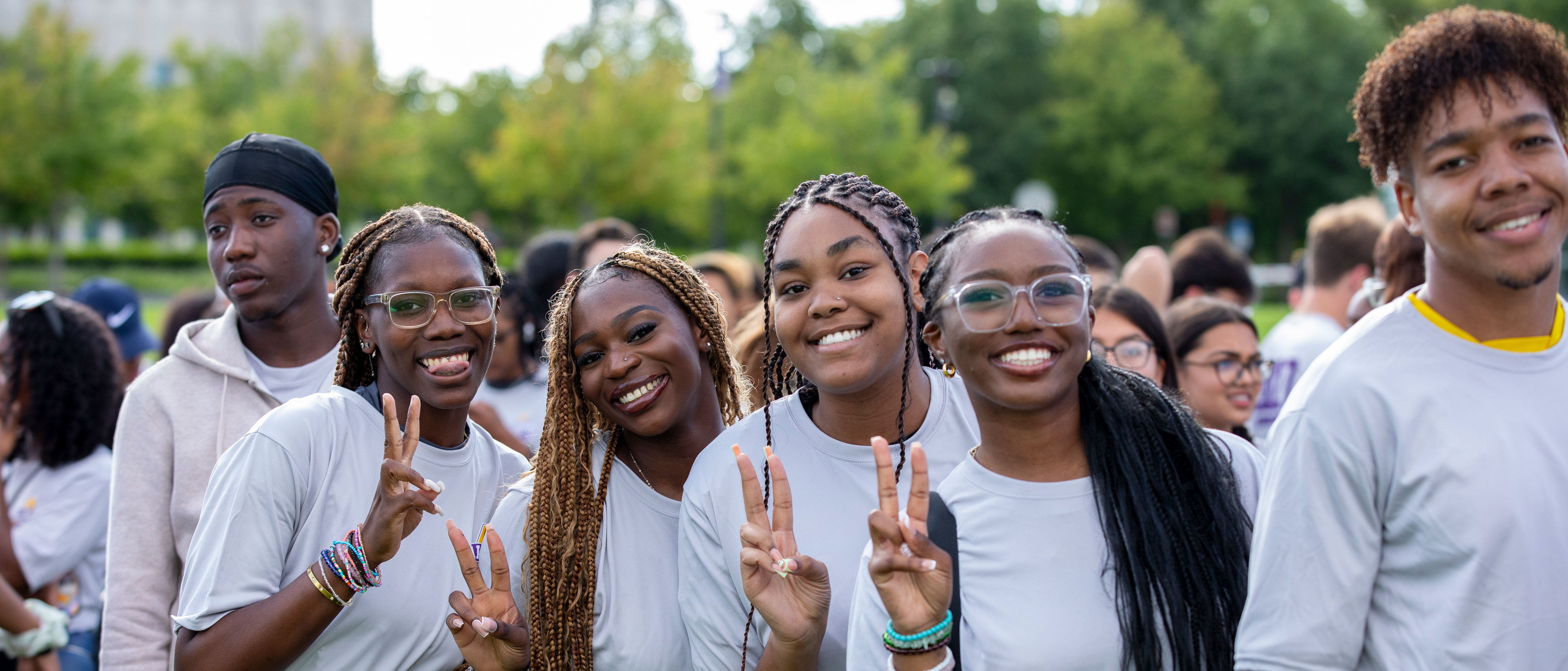 Six Black students wearing matching light gray t-shirts smile and pose for a photo. Four of the students hold up peace signs.