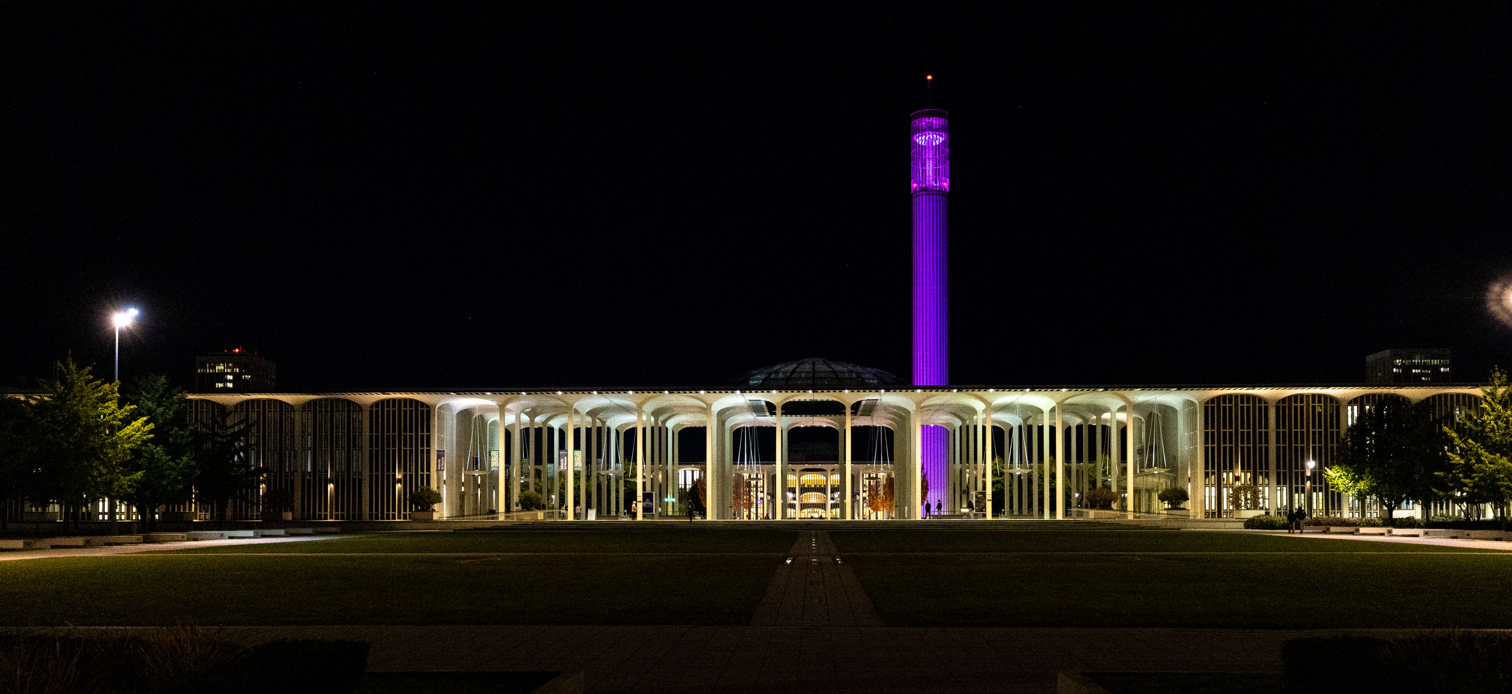 The UAlbany carillon tower lit up with purple lights and the Academic Podium buildings lit up with white lights at night.