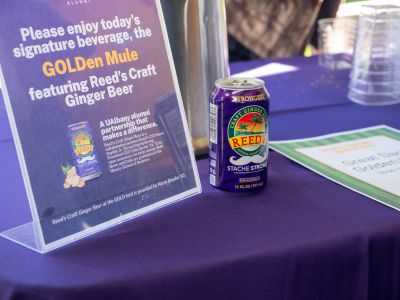 A purple can with a palm tree logo sits on a table next to a sign reading "Please enjoy today's signature beverage, the Golden Mule featuring Reed's Craft Ginger Beer."