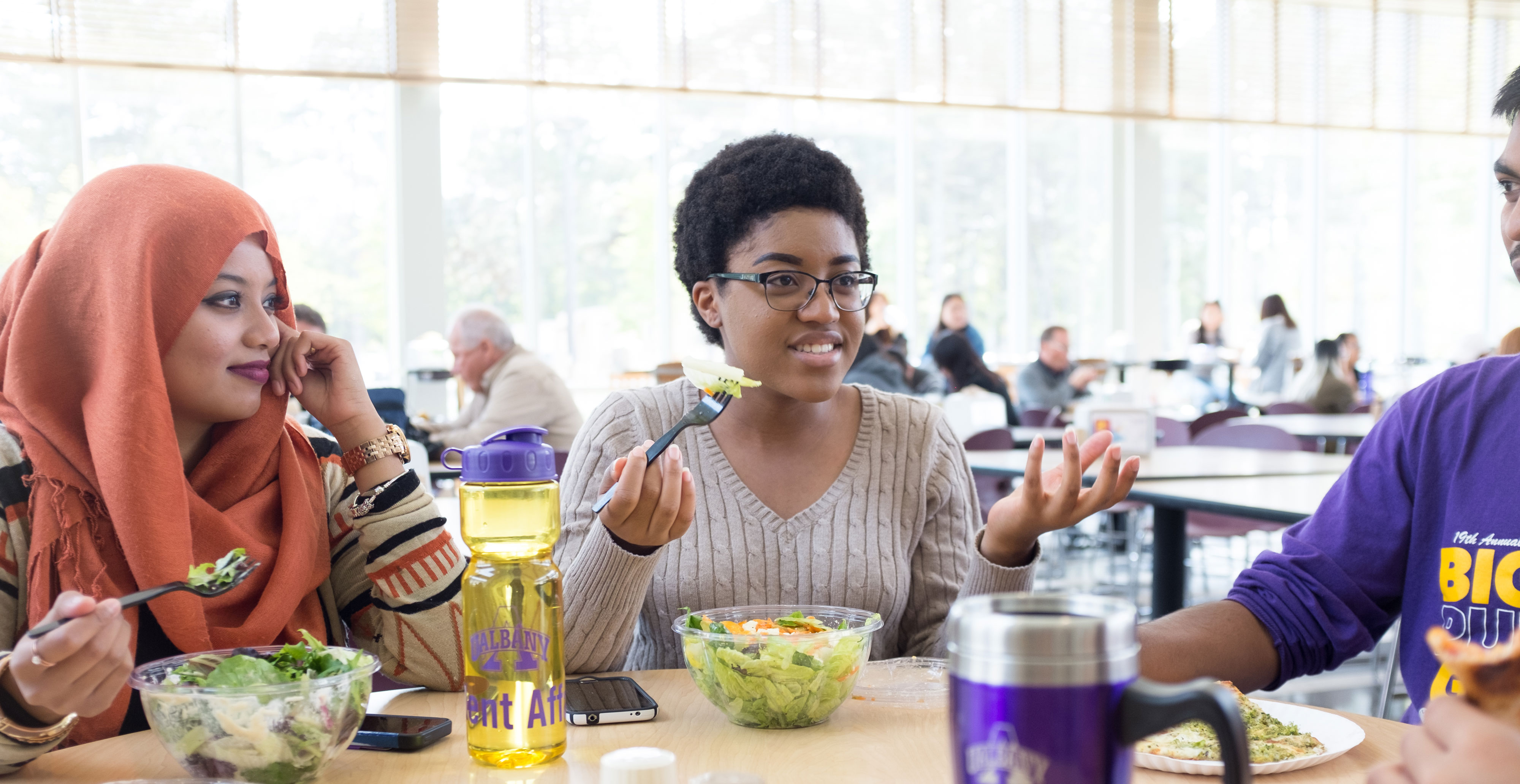 Three students gesture, smile and talk while sitting at a table eating salads.