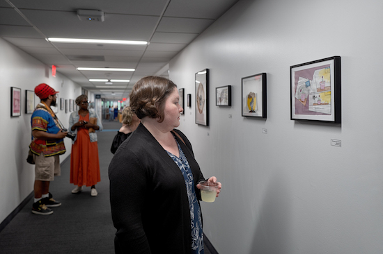 A woman with short brown hair in a black blazer holds a beverage in a plastic cup and looks at artwork hanging in a hallway as people mill about.