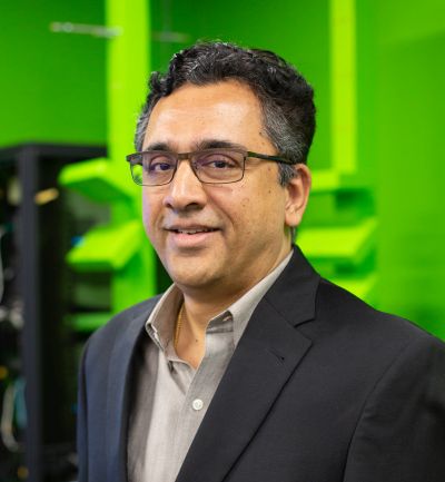 A portrait of a man smiling, wearing a dark dress jacket and eyeglasses against a bright green backgroun.