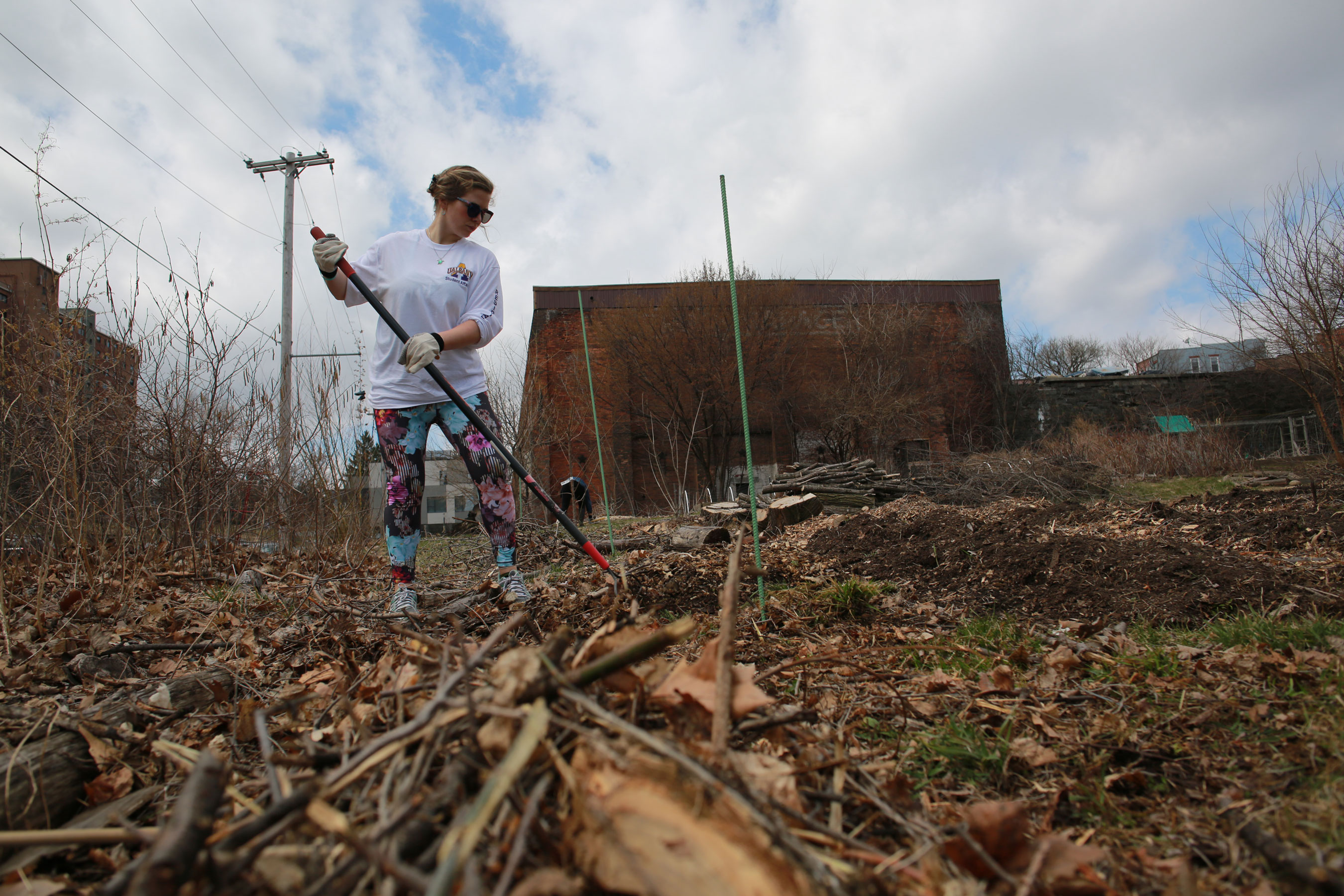 A student rakes debris in an empty urban lot on a cloudy day.