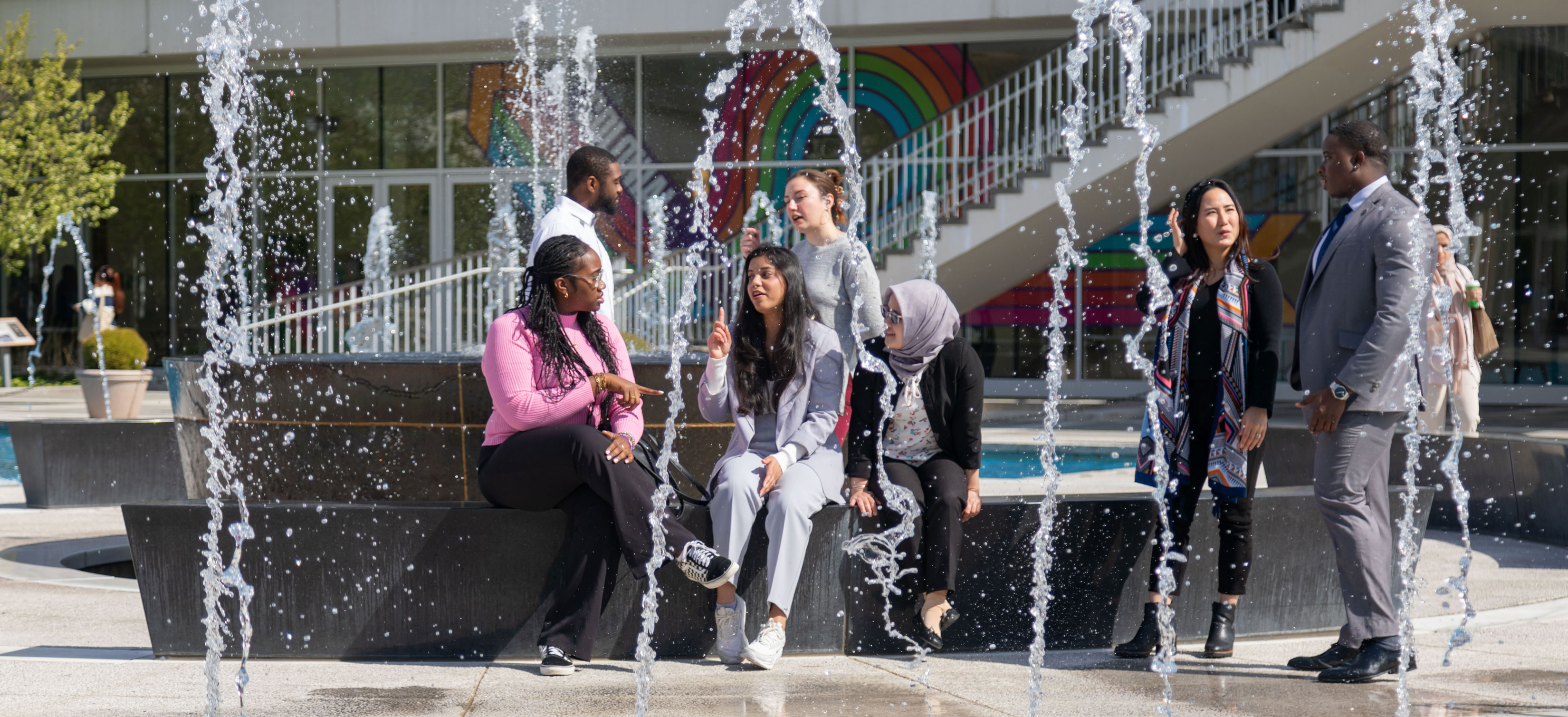 Seven student employees wearing business casual sit and stand around the main fountain on UAlbany's Uptown Campus.