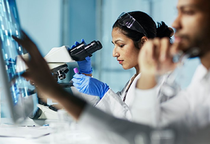 A student in a white coat uses a microscope in a UAlbany lab.
