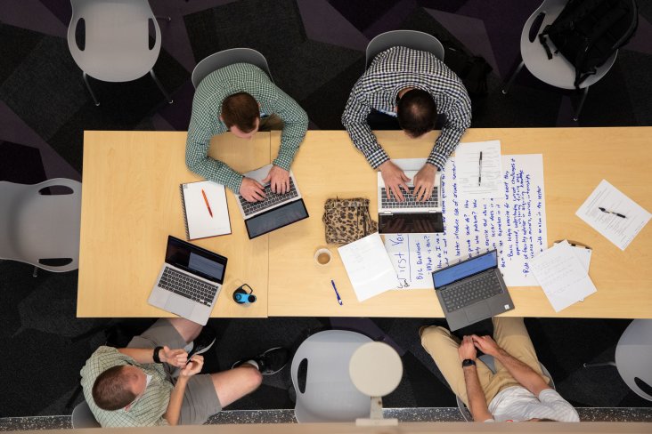 four men sitting at a table with their laptops open, and taking notes.