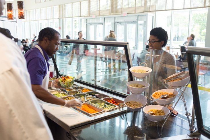 A dining employee prepares a salad for a student standing in front of the food station.