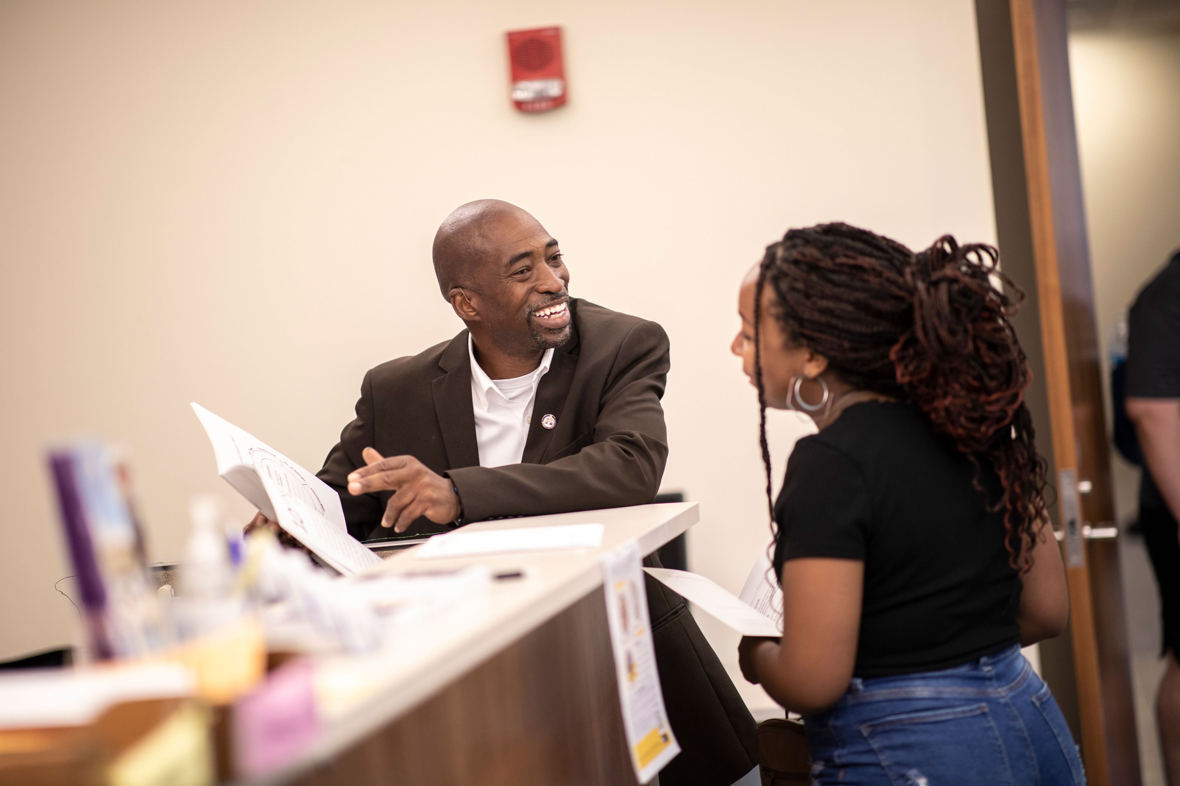 A staff member smiles as he speaks with a student inside an office. The student is looking at the pamphlet the staff member is pointing at.