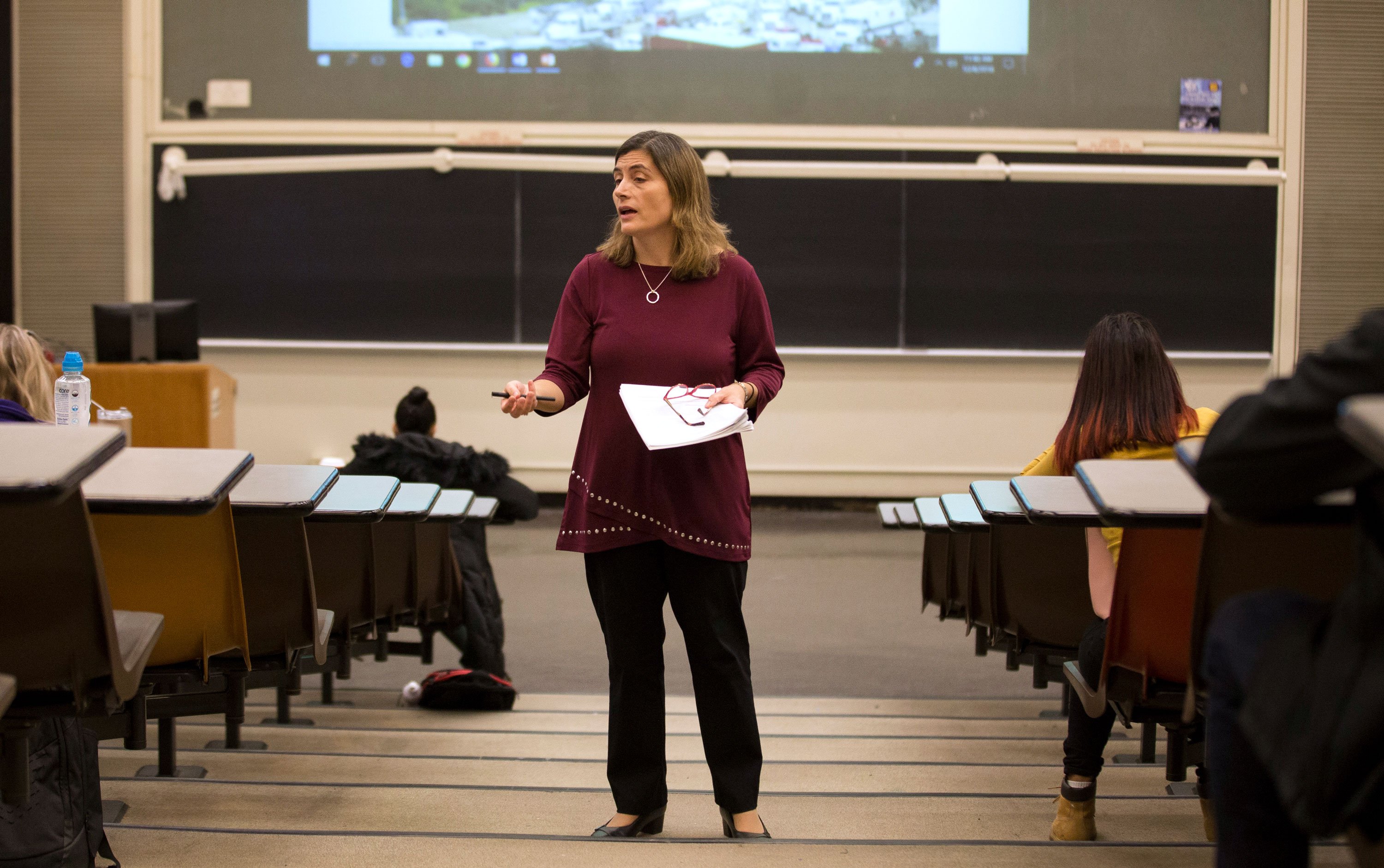 A faculty member holds a pen in one hand, and holds a stack of papers and eye glasses in the other hand, while teaching a class inside a large lecture hall.