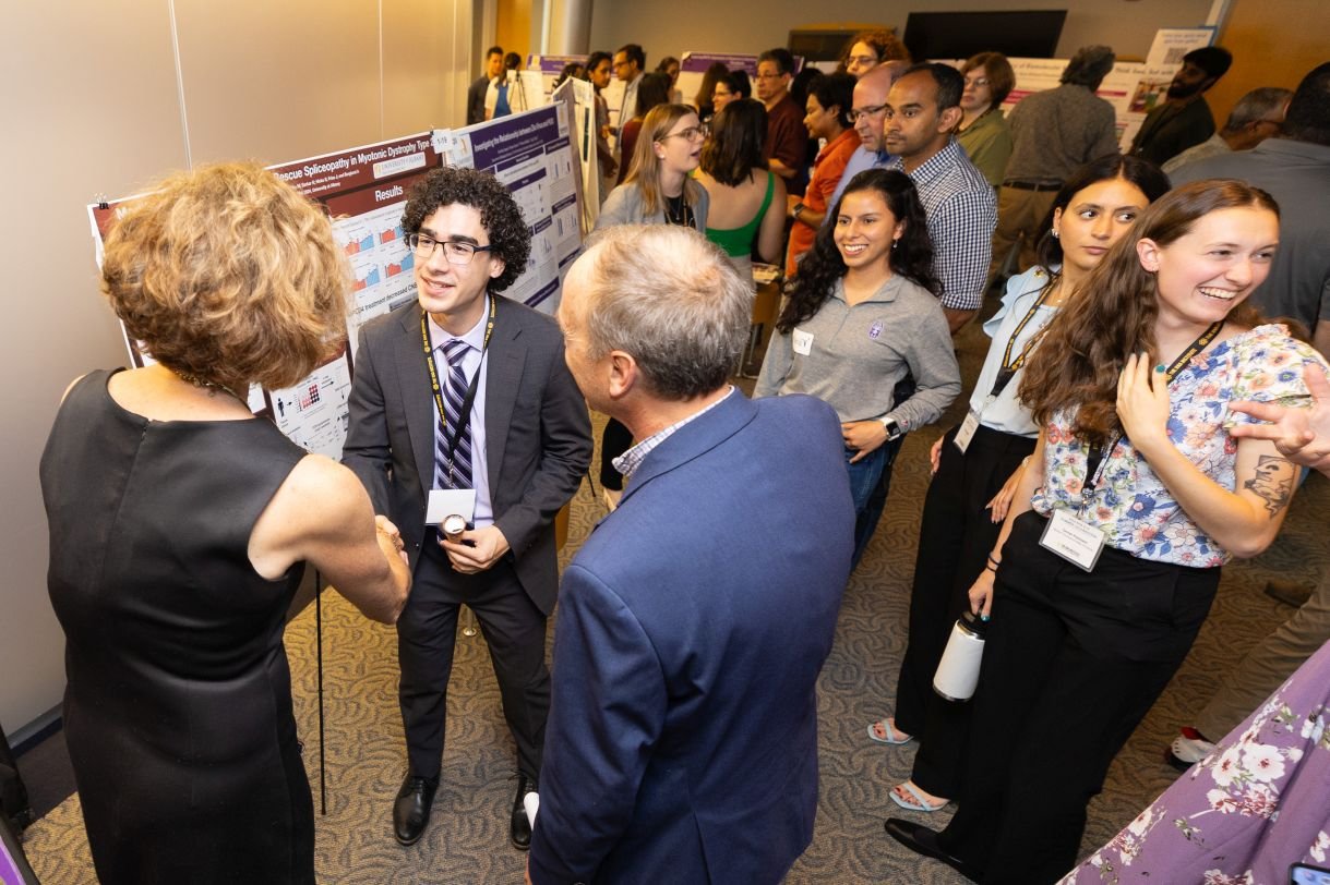 Students wearing nametags and business clothes stand with their posters in a crowded hall. 