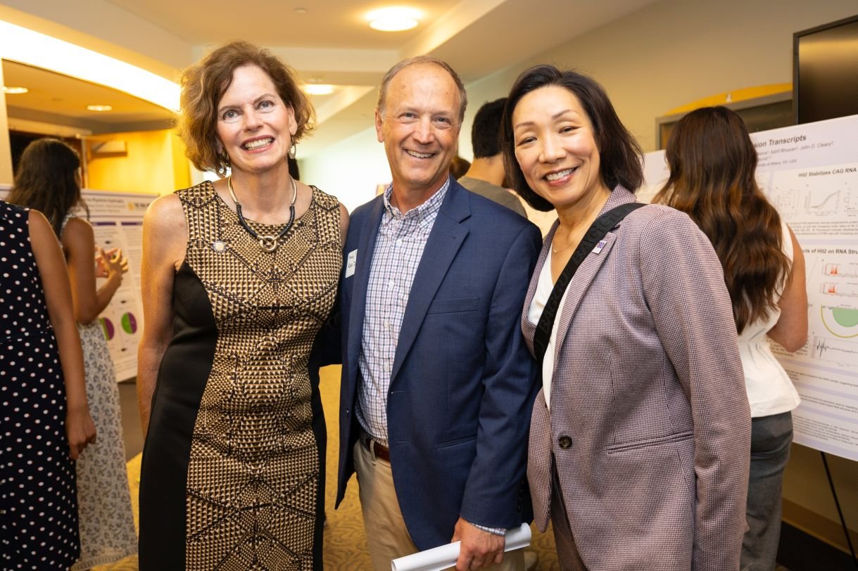 A woman in a gold and black dress, a man in a navy blazer, and a women in a lavender suit pose for a photo together. 