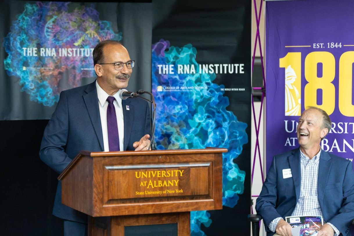 A man wearing a navy blazer, purple tie and glasses stands at a wooden podium that sys University at Albany. There are two colorful banners in the background that say “The RNA Institute” and one that says “180 University at Albany”.