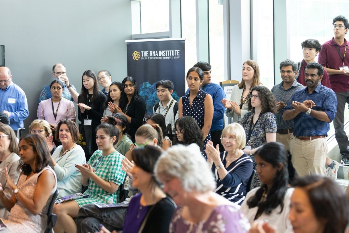 Image shows a bright sunlit room full of people listening to a man wearing a navy suit and purple tie. Many are smiling and applauding. 