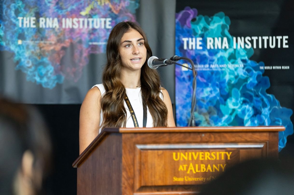 A young woman with long brown hair speaks into a microphone. Banners behind her say “The RNA Institute”.