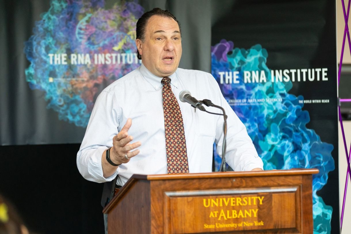 A man with brown hair, white button up shirt and red tie speaks into a microphone. Banners behind him say “The RNA Institute”.