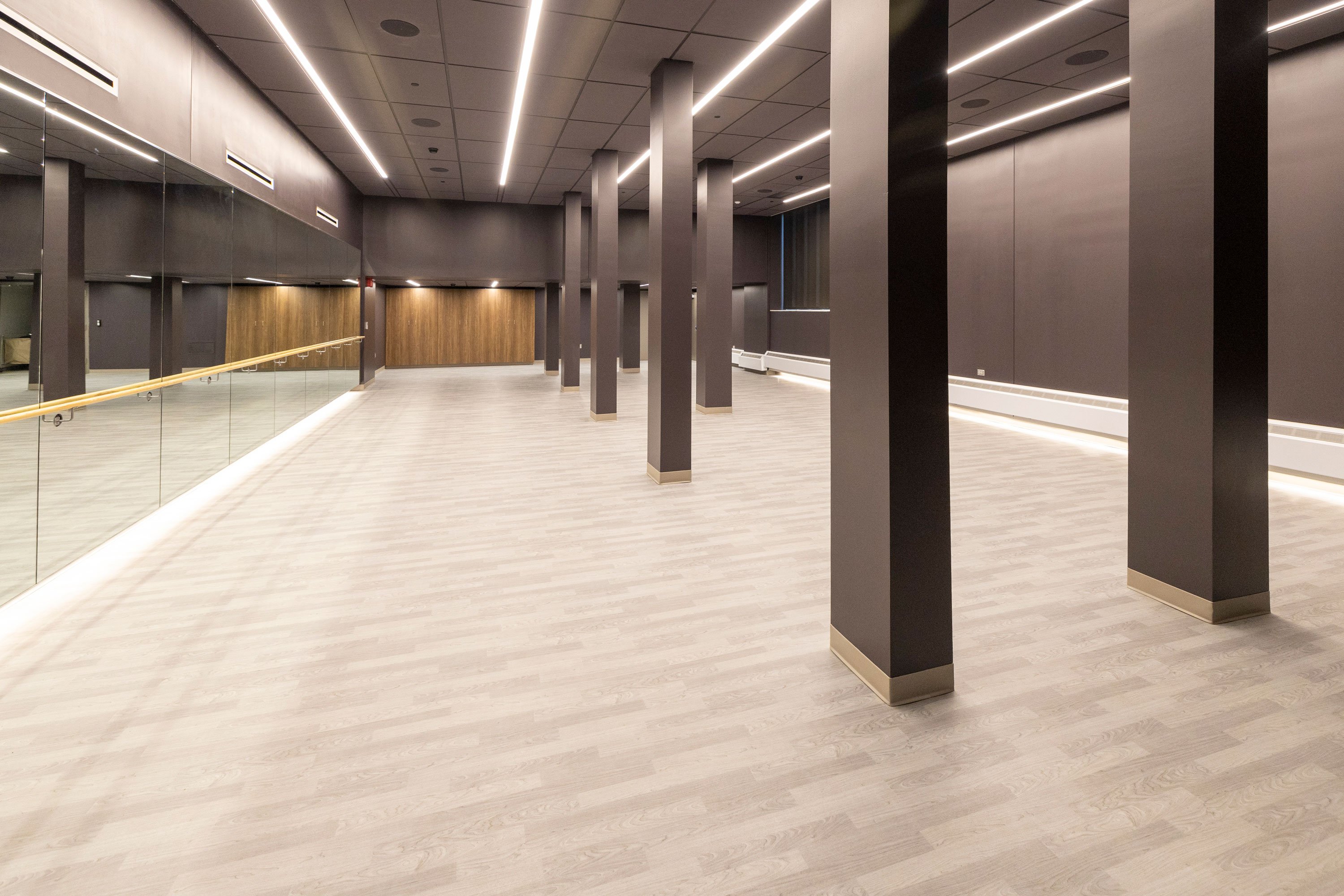 A group exercise room with a wood floor, a mirrored wall, a dance bar and gray columns.