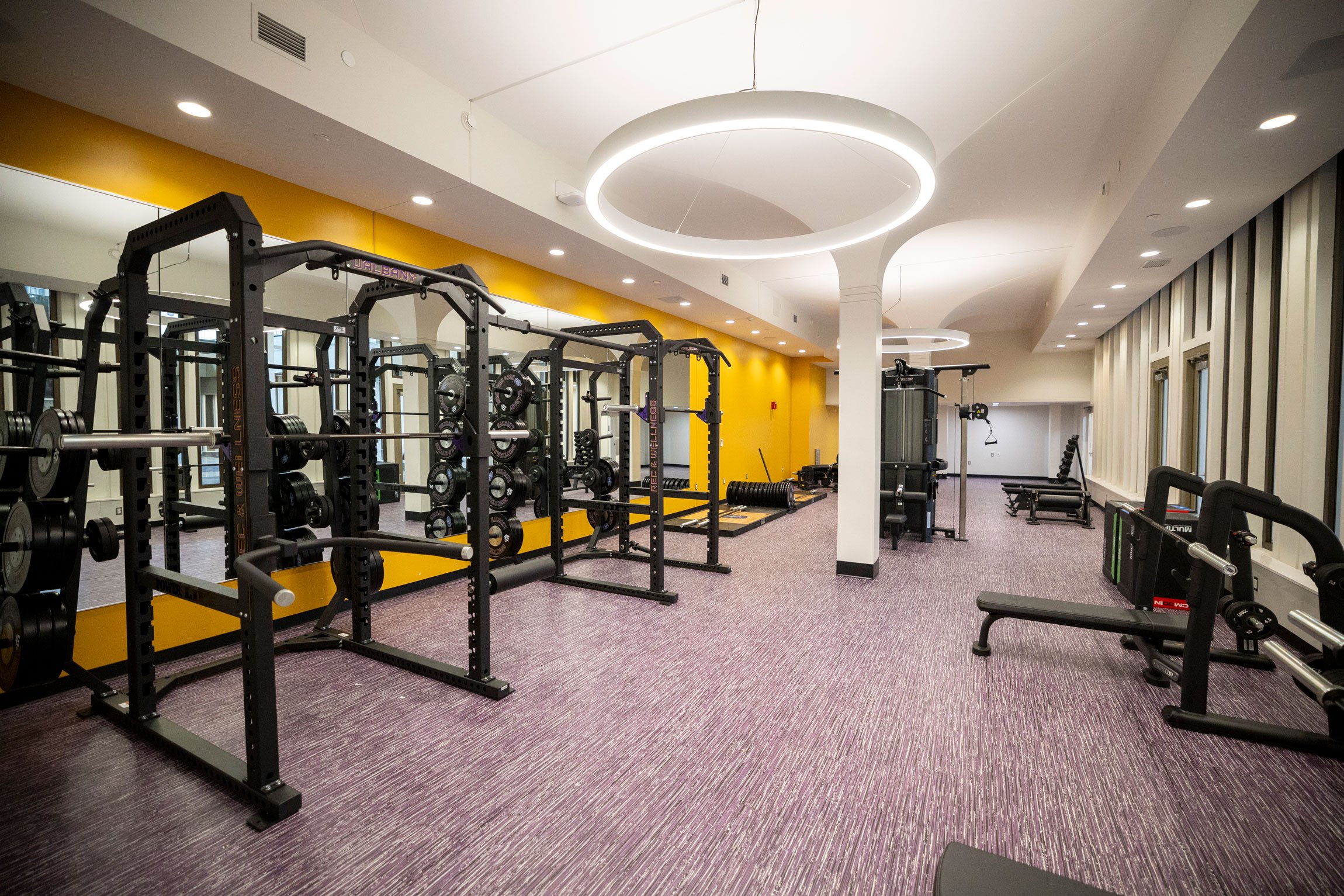 A weight room with various types of apparatus, purple carpet and a yellow wall.