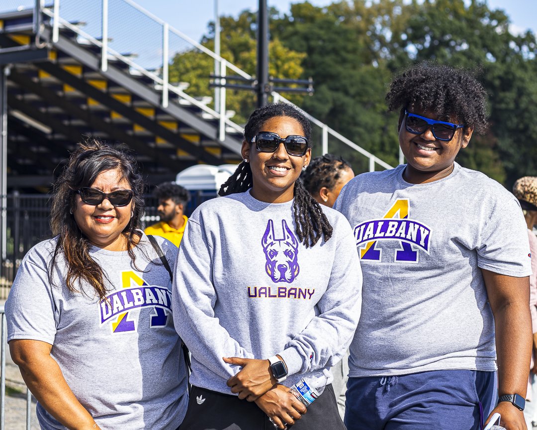 Three people wearing UAlbany t-shirts and sweatshirts smile and pose for a photo in front of stadium bleachers.