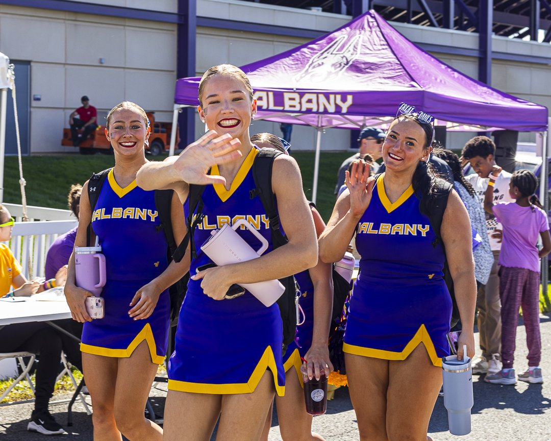 Three UAlbany cheerleaders smile and wave at the camera as they walk into the football stadium.