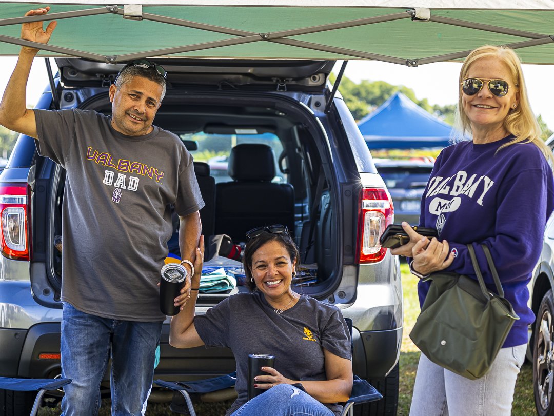 Two adults wearing UAlbany Mom and UAlbay Dad t-shirts stand beside a third seated adult, as they all smile and pose for a group photo.