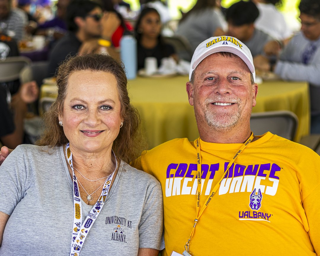 Two parents wearing UAlbany t-shirts smile for a photo while seated at a table for lunch.