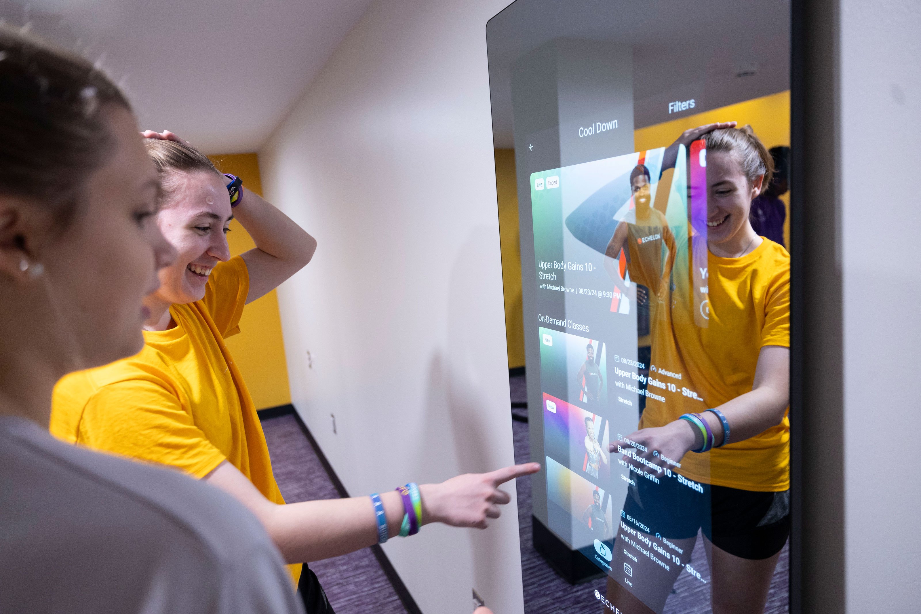 Two students smile as one of them uses a touch screen depicting cool down workout options.